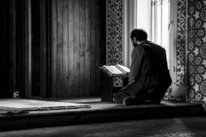 Uomo inginocchiato mentre legge un libro sacro in una stanza decorata con motivi islamici, illuminata dalla luce di una finestra.