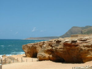 Paesaggio costiero con imponenti scogliere di roccia arenaria dorata, grotte naturali, una piccola spiaggia sabbiosa di El-Haouria e il mare agitato sotto un cielo blu.