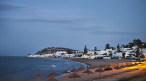 Spiaggia di Gammarth al tramonto con ombrelloni in paglia, mare calmo e case bianche lungo la costa tunisina.