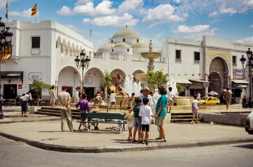 Piazza centrale nella medina di Tunisi con fontana, bambini che giocano e edifici bianchi storici sotto il cielo azzurro.