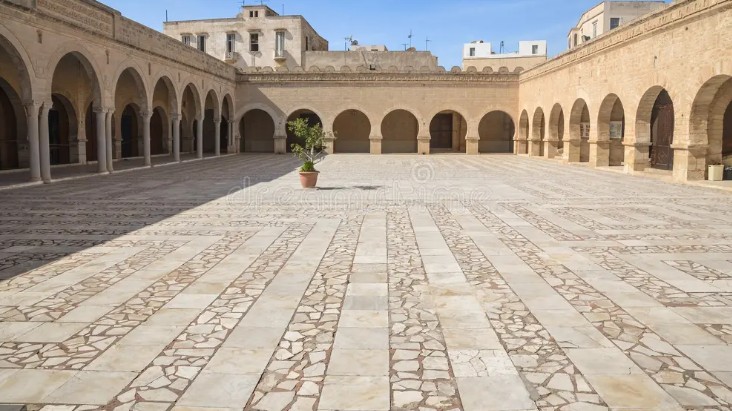 Cortile interno pavimentato della Grande Moschea di Sousse, Tunisia, circondato da portici ad archi, con un albero in vaso sullo sfondo e cielo sereno.