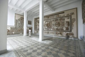 Sala museo luminosa con colonne, mosaici romani alle pareti e pavimento a mosaico; una persona osserva l’esposizione